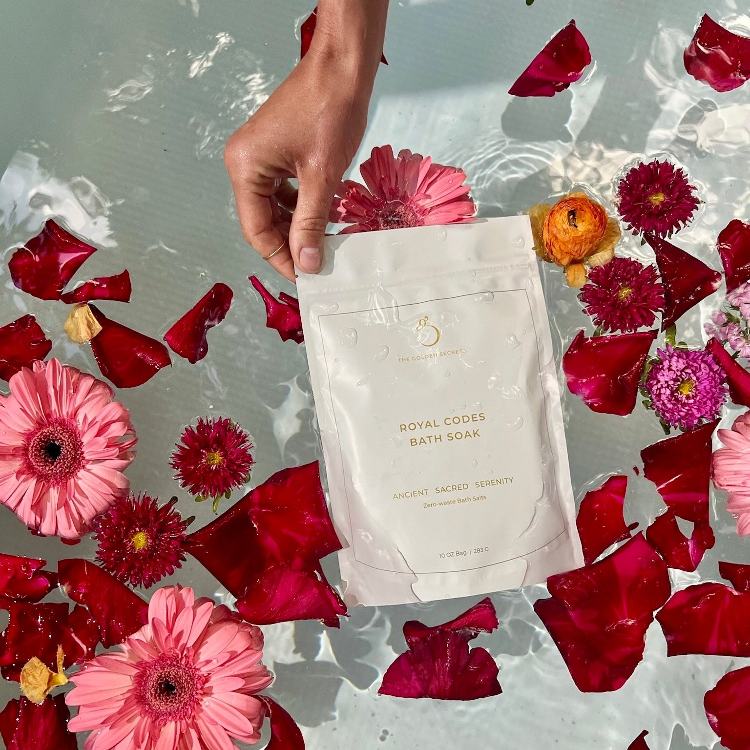 A hand holds a pouch labeled Royal Codes Bath Soak from THE GOLDEN SECRETS above water filled with floating pink flowers, red petals, and an orange flower, showcasing this mineral-rich bath soak infused with pure Zechstein Magnesium Chloride.