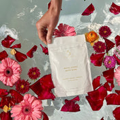 A hand holds a pouch labeled Royal Codes Bath Soak from THE GOLDEN SECRETS above water filled with floating pink flowers, red petals, and an orange flower, showcasing this mineral-rich bath soak infused with pure Zechstein Magnesium Chloride.
