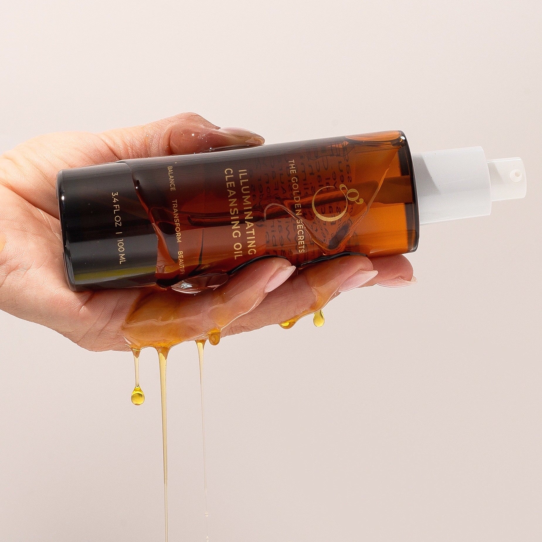 A hand holding a bottle of Javobi Illuminating Cleansing Oil, with golden oil dripping from the bottom against a solid light beige background.