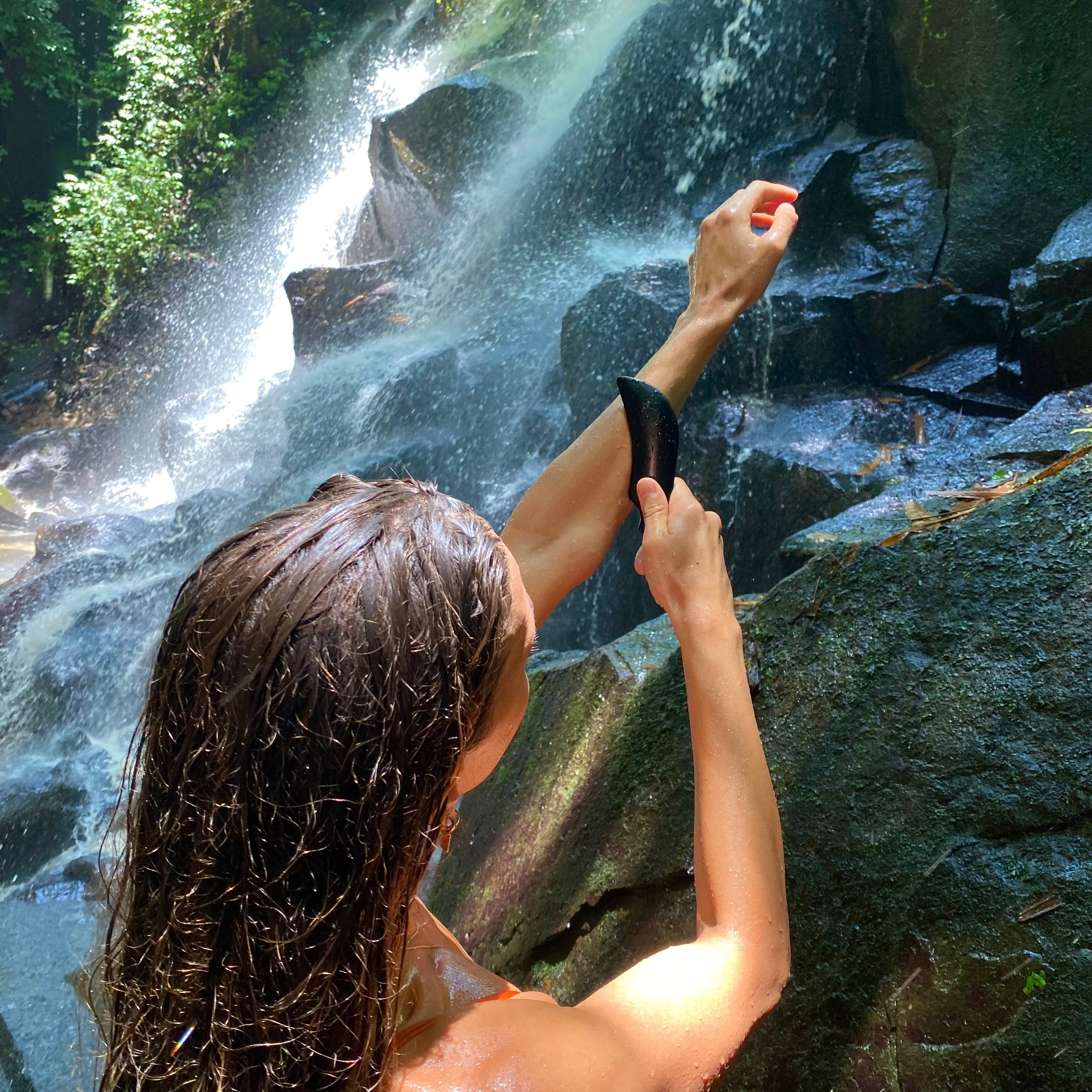 A woman with wet brown hair stands at a waterfall, wearing a black wrist strap or band around her wrist, with mossy rocks and green foliage in the background.