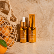 The Dynamic Duo—two gold bottles—rests on a beige stone beside a net bag of oranges, creating the perfect pair for radiant, nourished skin.