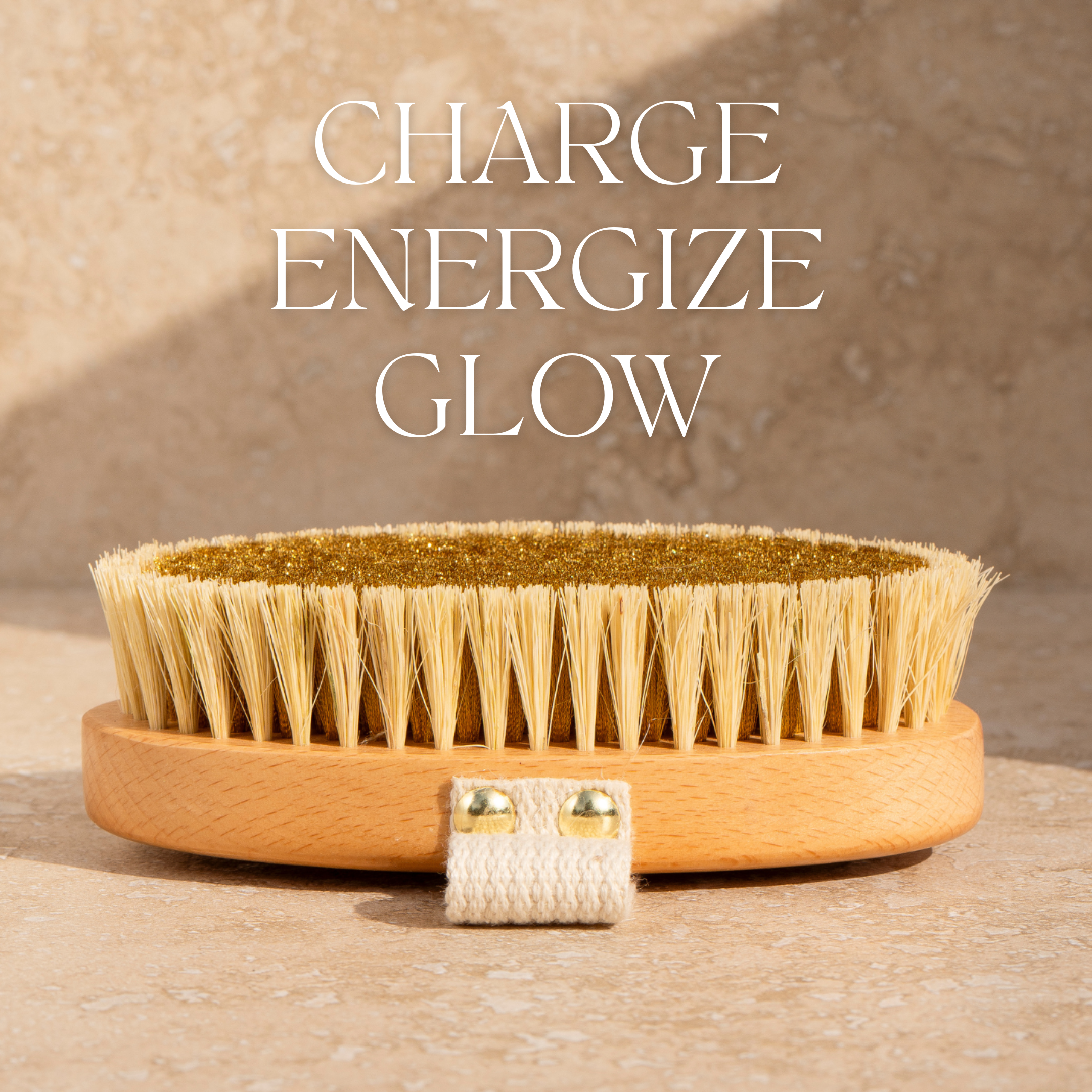 On a beige surface, the Ionic Copper Dry Brush from THE GOLDEN SECRETS—crafted with natural bristles and ion-charged copper bristles—rests in sunlight, casting a shadow. Above the brush, “CHARGE ENERGIZE GLOW” is displayed in white text. Designed to support lymphatic flow, this brush gently exfoliates dead skin cells for radiant skin.