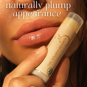 A close-up of a person’s glossy lips holding a tube of THE GOLDEN SECRETS Organic Coconut Lip Balm. Text above reads: naturally plump appearance—perfect to hydrate lips all day.