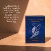 A box of THE GOLDEN SECRETS The Golden Secrets Guidance Cards rests on a sunlit surface. Next to it, text reads: “Each moment with The Golden Secrets Guidance Cards by THE GOLDEN SECRETS is an invitation to slow down, tune in, and listen more deeply to your inner knowing—offering comfort and peace along the way.”.