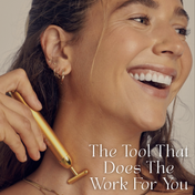 A smiling person glides THE GOLDEN SECRETS Gold Pulse Firming Face Wand, plated in 24k gold, over their cheek, enjoying its soothing touch. Text on the image reads, "The Tool That Does The Work For You," showcasing how it lifts and sculpts skin for a rejuvenated glow.