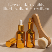 Two golden bottles from THE GOLDEN SECRETS Lymph & Lift Kit stand on a beige surface next to a facial dry brushing brush. Text above reads, Leaves skin visibly lifted, radiant & resilient.