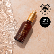 A brown bottle of THE GOLDEN SECRETS Golden Hour Duo body oil with a gold cap rests on a wet, beige surface. Infused with nourishing oils for radiant skin, the bottle is surrounded by water ripples, and two certification badges—EWG Verified and Think Dirty Top Summer Pick—are displayed nearby.