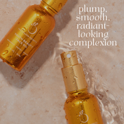 Two gold bottles of THE GOLDEN SECRETS Dynamic Duo face oil rest on a wet beige surface, with water flowing around one bottle. Text reads: plump, smooth, radiant-looking complexion.