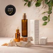 THE GOLDEN SECRETS Dynamic Duo—a hydrating mist and face oil—are elegantly arranged on beige stones, accented by fresh green leaves. A Think Dirty Verified badge appears in the upper left corner, while a textured, softly lit background enhances the nourishing beauty of this signature skincare set presented alongside a white box.