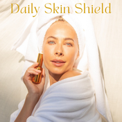 A woman in a white robe and towel holds THE GOLDEN SECRETS Dynamic Duo bottle near her face. Sunlight highlights her clear skin, radiant from her favorite hydrating mist. The text Dynamic Duo appears above her in gold letters.