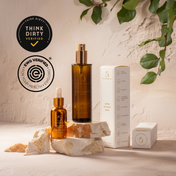 The Ultimate Glow Trio Kit by THE GOLDEN SECRETS features brown glass skincare bottles and a white box arranged on rock pieces, promising to hydrate, nourish, and lift your skin. Green leaves hang in the top right corner, while “Think Dirty Verified” and “EWG Verified” badges are displayed on the left side of the image.