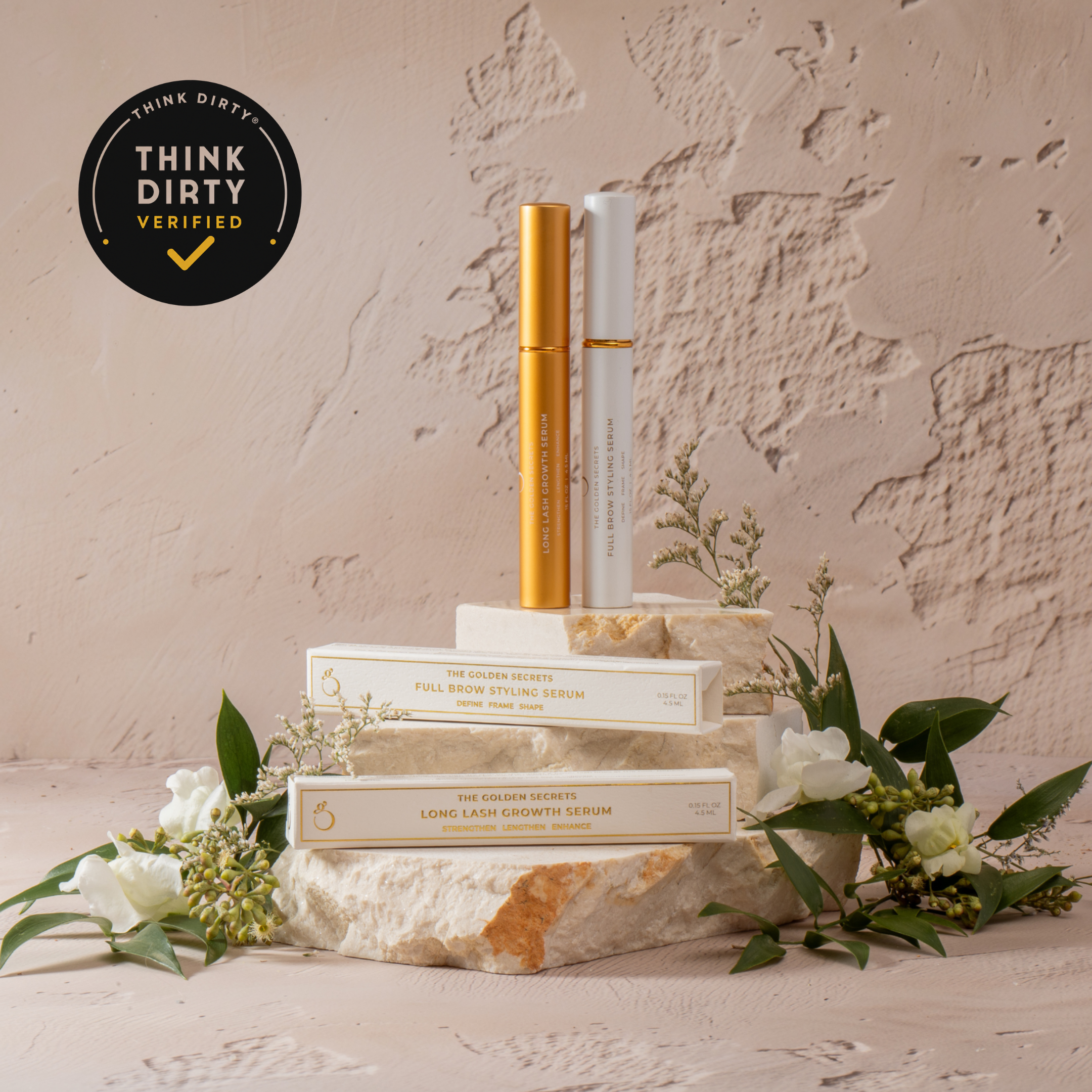 THE GOLDEN SECRETS Lash & Brow Duo—featuring two gold and white beauty serums—is elegantly displayed on stacked boxes amid white flowers. Against a textured beige background, a black Think Dirty Verified badge appears in the top left corner, spotlighting the triple peptides formula designed for fuller-looking lashes.