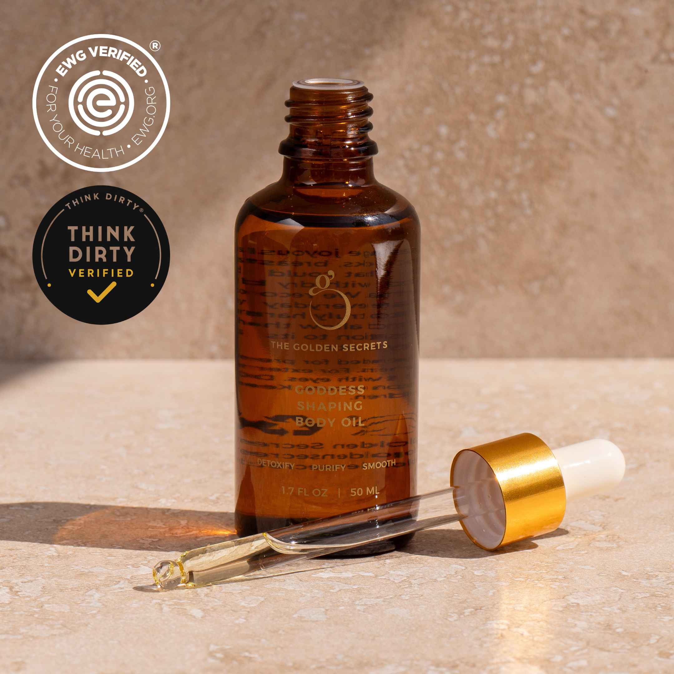 A dropper-topped amber bottle of THE GOLDEN SECRETS Body Sculpting Kit Body Oil sits on a beige surface, making it an ideal complement to your body care routine. EWG VERIFIED and THINK DIRTY VERIFIED logos are displayed in the top left corner.