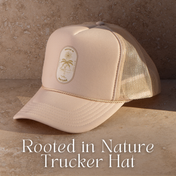 A beige trucker hat from THE GOLDEN SECRETS, featuring a palm tree patch and the words “Rooted in Nature,” rests on a stone surface, evoking the tranquility of golden hour. Text below reads: The Golden Secrets Trucker Hats.