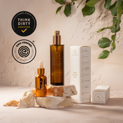 THE GOLDEN SECRETS Sacred Triad mist and serum bottles with their product box displayed on stone, surrounded by green leaves. Think Dirty and EWG Verified certification logos are shown, highlighting these clean, health-conscious beauty products designed to elevate your glow ritual and promote a radiant complexion.