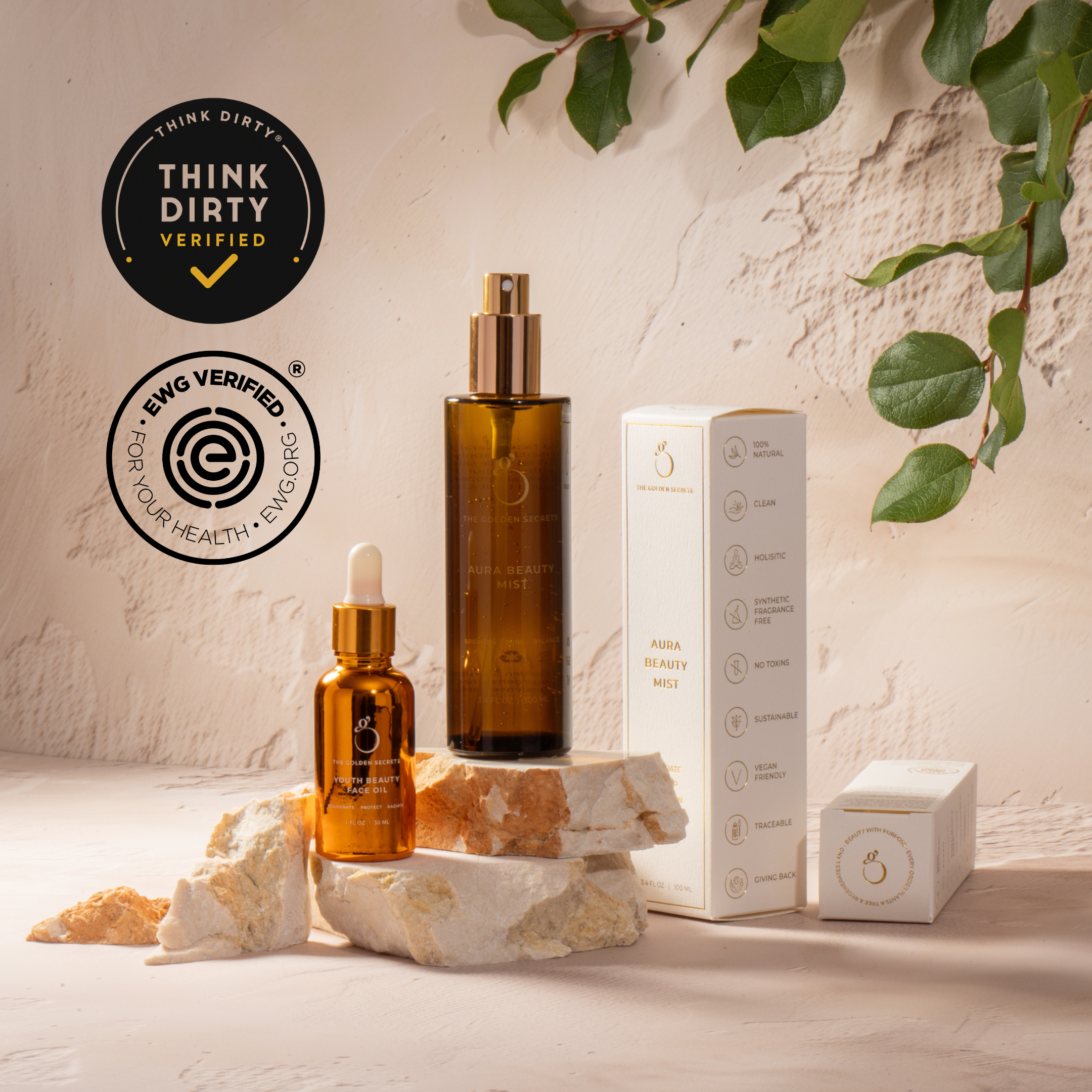THE GOLDEN SECRETS Sacred Triad mist and serum bottles with their product box displayed on stone, surrounded by green leaves. Think Dirty and EWG Verified certification logos are shown, highlighting these clean, health-conscious beauty products designed to elevate your glow ritual and promote a radiant complexion.