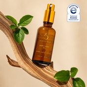 A brown bottle of THE GOLDEN SECRETS Mom & Baby Oil with a gold pump rests on a curved tree branch, nestled among vibrant green leaves. Expertly formulated to nurture baby’s delicate skin barrier, this nourishing oil is gentle enough for little ones and soothing for mom’s changing skin. A National Eczema Association Accepted badge appears in the top right corner.