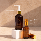 Two brown glass bottles of THE GOLDEN SECRETS Mom & Baby skincare oil—one larger with a pump labeled “For Mama” and one smaller with a spray labeled “For Baby”—are displayed on a white block against a beige background. This nourishing duo is thoughtfully crafted to support mom’s changing skin and protect your baby’s delicate barrier.