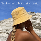 A woman with a blonde braid wears the Golden Sun Bucket Hat and faces rocky cliffs under a blue sky. White text above reads, "Seek shade... but make it cute.