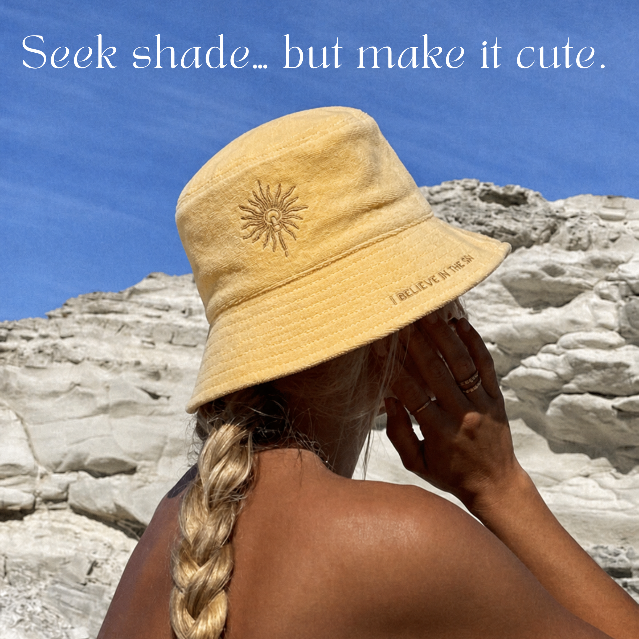 A woman with a blonde braid wears the Golden Sun Bucket Hat and faces rocky cliffs under a blue sky. White text above reads, "Seek shade... but make it cute.