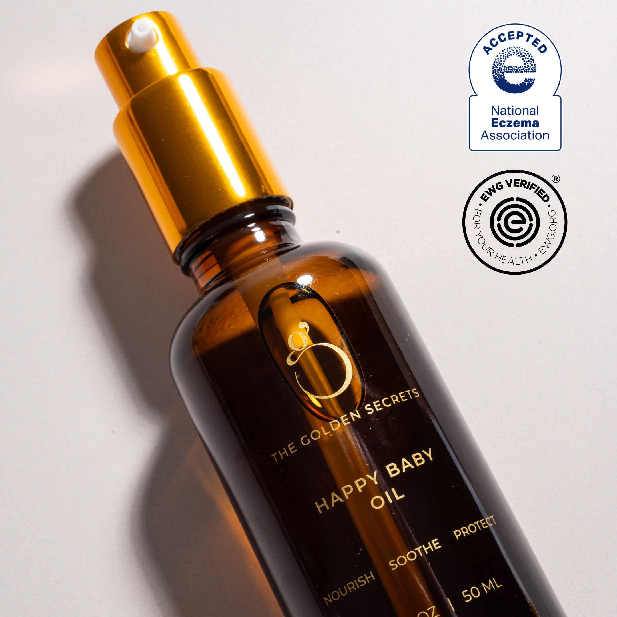 An amber bottle labeled "Happy Baby Oil" by THE GOLDEN SECRETS features a gold spray top. It displays two seals: "Accepted National Eczema Association" and "EWG Verified." The label reads "nourish, soothe, protect." This organic baby oil is expertly formulated for sensitive skin to provide gentle care and relieve dryness.