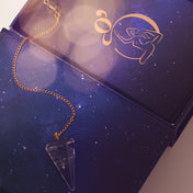 A clear crystal tarot card pendant necklace with a gold chain, displayed on a dark blue starry box with a gold card design.