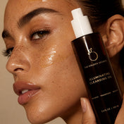 A woman with glowing, freckled skin holds a bottle labeled Cleansing Duo by THE GOLDEN SECRETS close to her face. A streak of the cleansing oil is visible on her cheek, emphasizing her radiant complexion.