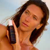 A woman outdoors holds a dark bottle with a white cap, likely a skincare or cosmetic product. The bottle has gold lettering that appears to read 'SKIN1000' and 'MORNING AFTER.