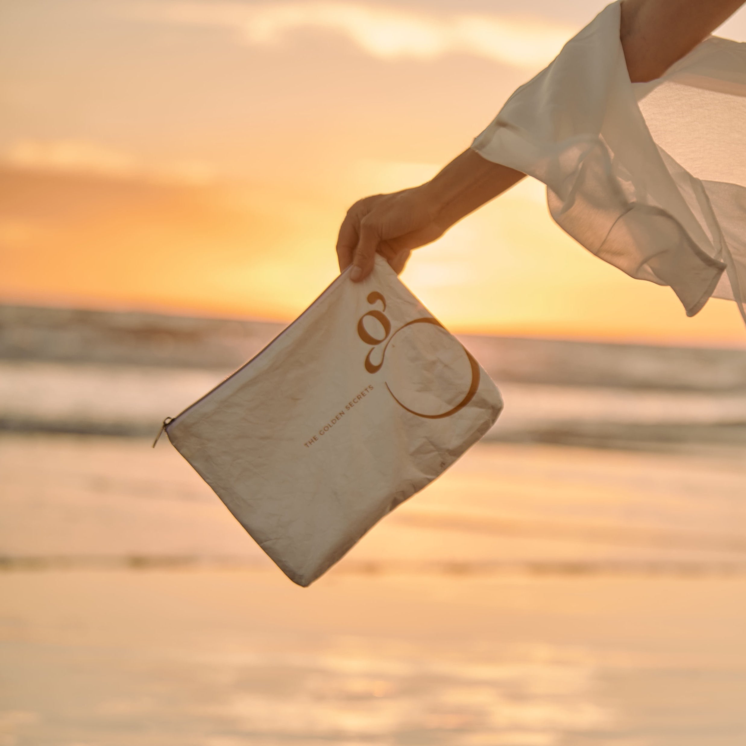 A hand in a flowing sleeve holds The Golden Secrets Skincare Bags by THE GOLDEN SECRETS—a beige, water-resistant pouch with gold lettering—perfect for organizing skincare and travel essentials—set against a beach sunset backdrop with waves and a glowing sky.