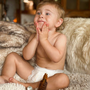 Naked baby sitting on a fluffy surface with a bottle of baby oil nearby.