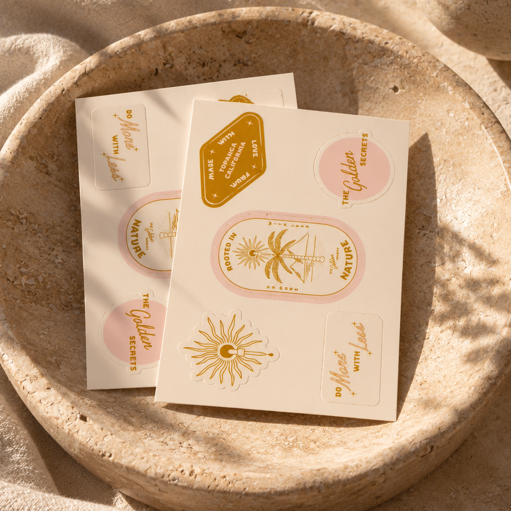 Two sheets from THE GOLDEN SECRETS Golden Sun Sticker Pack, a limited edition collection featuring gold and pink stickers with sun motifs, palm trees, and uplifting phrases, rest inside a round beige stone bowl atop a textured surface bathed in warm sunlight—evoking sustainable style with every detail.
