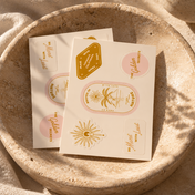 Two sheets from THE GOLDEN SECRETS Golden Sun Sticker Pack, a limited edition collection featuring gold and pink stickers with sun motifs, palm trees, and uplifting phrases, rest inside a round beige stone bowl atop a textured surface bathed in warm sunlight—evoking sustainable style with every detail.