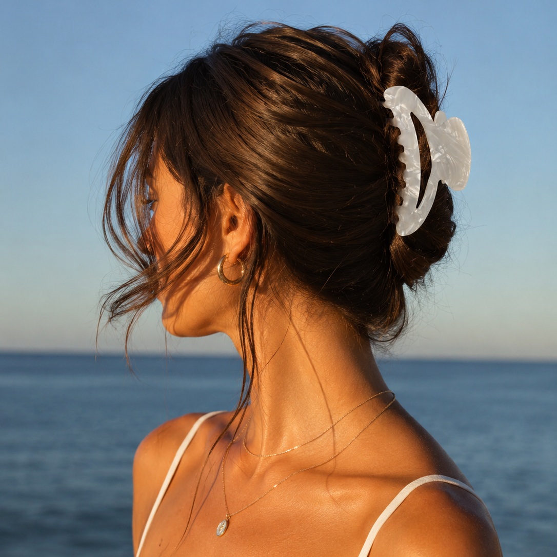 A woman with brown hair secures her locks with THE GOLDEN SECRETS Goddess Hair Clip as she gazes out at the ocean. Dressed in a thin-strap top, gold necklace, and hoop earring, the sunlight accentuates her radiant skin and glossy hair—an effortless reflection of her self-care beauty rituals.