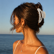 A woman with brown hair secures her locks with THE GOLDEN SECRETS Goddess Hair Clip as she gazes out at the ocean. Dressed in a thin-strap top, gold necklace, and hoop earring, the sunlight accentuates her radiant skin and glossy hair—an effortless reflection of her self-care beauty rituals.