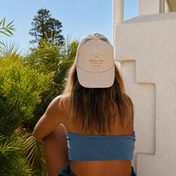Wearing THE GOLDEN SECRETS’ The Golden Secrets Trucker Hat and a blue strapless top, a person sits with their back to the camera next to lush greenery and a white building, soaking up golden hour sunlight.