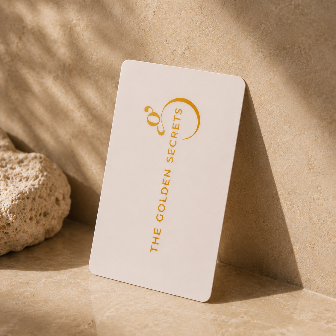 A white digital The Golden Gift Card from THE GOLDEN SECRETS, adorned with gold lettering, rests against a textured beige stone and casts a gentle shadow on a marble surface illuminated by warm natural light—an elegant choice for email delivery.