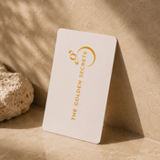 A white digital The Golden Gift Card from THE GOLDEN SECRETS, adorned with gold lettering, rests against a textured beige stone and casts a gentle shadow on a marble surface illuminated by warm natural light—an elegant choice for email delivery.