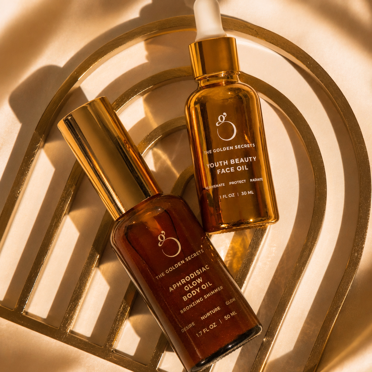 Two amber bottles with gold caps rest on a gold decorative tray, perfectly embodying the Golden Hour Duo from THE GOLDEN SECRETS. One bottle is Aphrodisiac Body Oil, while the other is Youth Beauty Face Oil with a dropper—both award-winning formulas crafted to nourish and enhance radiant skin. Sunlight casts soft shadows around them, emphasizing their luxurious allure.