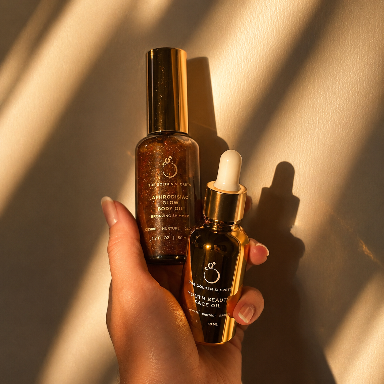 A hand holds two gold bottles—the Golden Hour Duo by THE GOLDEN SECRETS: Aphrodisiac Body Oil with a spray top, and Youth Beauty Face Oil with a dropper—both reflecting warm sunlight on a beige background for naturally radiant skin.