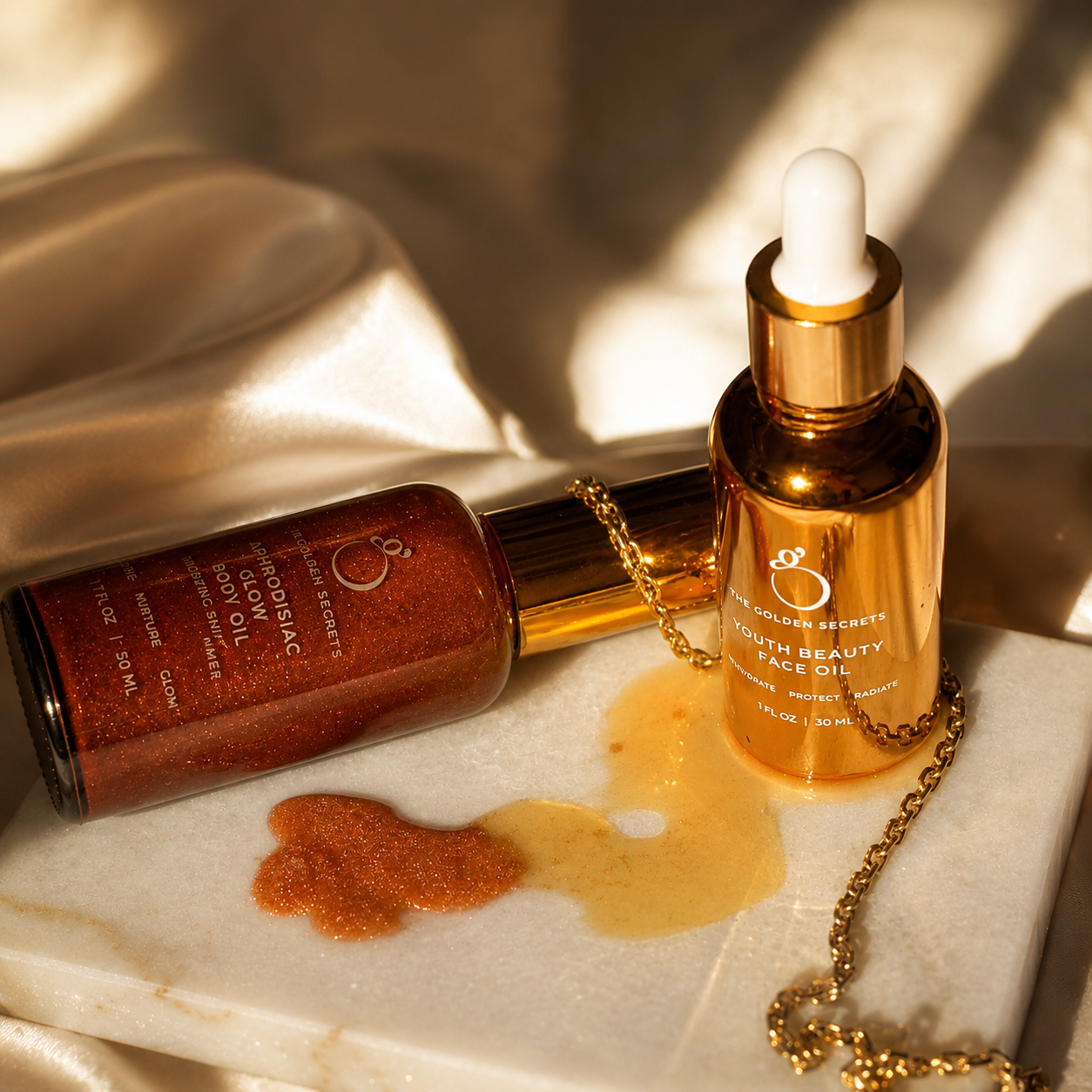 Two gold bottles of THE GOLDEN SECRETS' Golden Hour Duo beauty oil rest atop a marble surface—one elegantly tipped, letting its luminous oil spill out—while a delicate gold chain lies nearby. Against a backdrop of silky, cream-colored fabric illuminated by warm light, this luxurious scene showcases the Golden Hour Duo's promise of radiant skin.