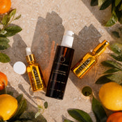 Three skincare bottles from The Golden Secrets—a face oil, a cleansing oil, and a face mist—surrounded by fresh lemons, oranges, and leaves on a stone surface.