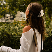 A woman with wavy brown hair, dressed in a white off-shoulder top and wearing THE GOLDEN SECRETS Goddess Hair Clip, stands outdoors near lush green bushes, her radiant skin suggesting dedicated self-care beauty routines.