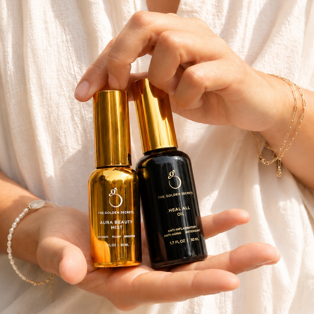 A person in a white outfit holds two skincare bottles—the gold-labeled Aura Beauty Mist and the black-labeled Heal All Oil, both part of THE GOLDEN SECRETS Hydrate & Heal Duo—formulated to support your skin barrier and microbiome.