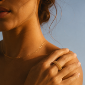 A woman wearing a delicate gold necklace and THE GOLDEN SECRETS Gold Reflexology Acupressure Massage Ring on her hand gently touches her collarbone, with warm sunlight casting soft shadows on her skin against a pale blue background.