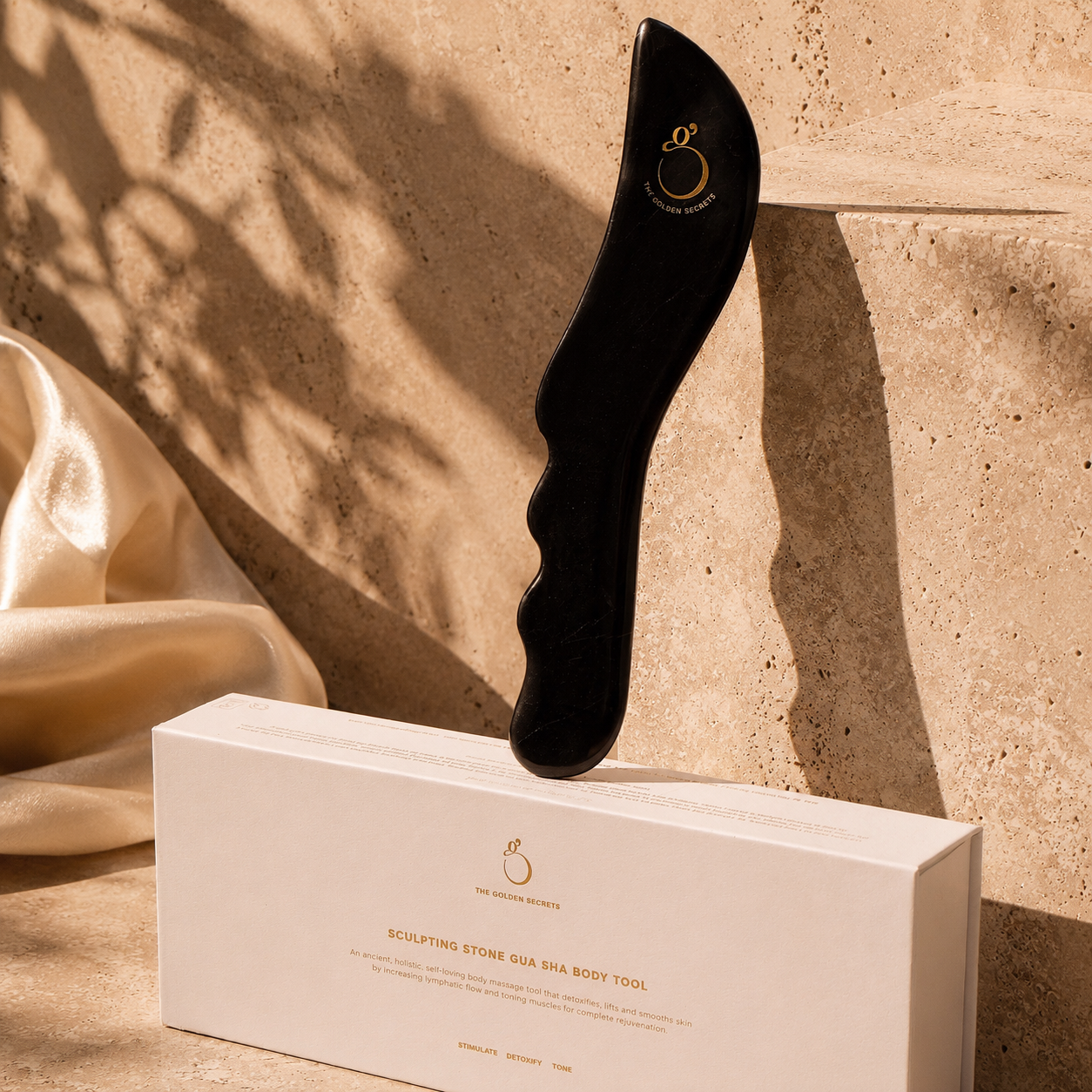 The Sculpting Stone Gua Sha Body Tool by THE GOLDEN SECRETS, crafted from a premium body sculpting stone in black, stands upright on its white packaging box, set against a backdrop of beige stone steps and soft fabric.