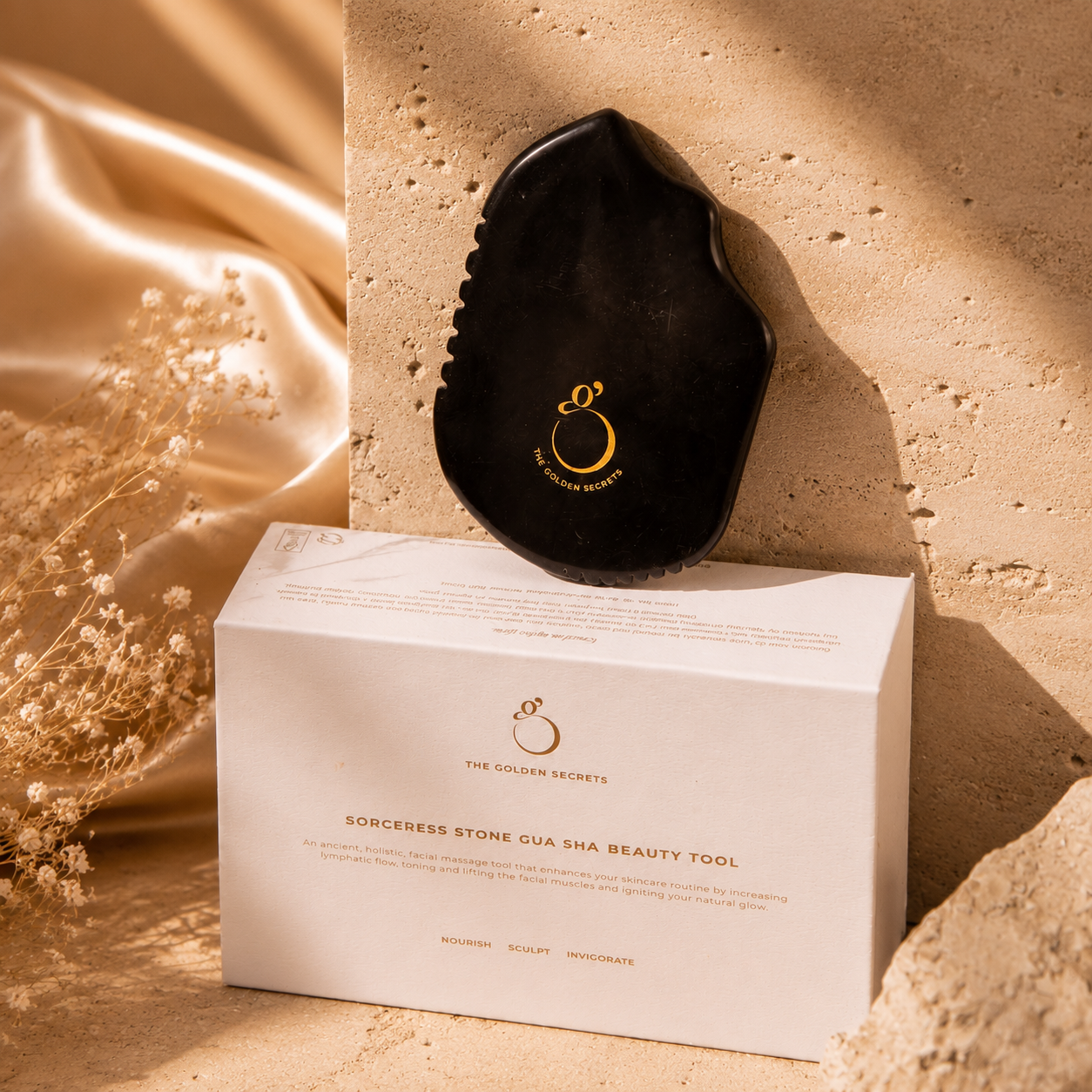 A black Sorceress Stone Gua Sha Beauty Tool by THE GOLDEN SECRETS, featuring a gold logo and crafted for effective facial sculpting, rests against a textured stone beside a white box labeled with its product name. Dried flowers and satin fabric complete the background.