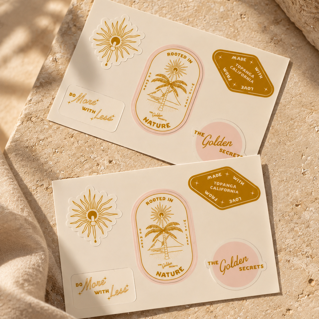 Two sheets from THE GOLDEN SECRETS’ Golden Sun Sticker Pack lie on a sandy beige surface. Showcasing radiant suns, palm trees, and uplifting phrases like ROOTED IN NATURE, DO MORE WITH LESS, and THE GOLDEN SECRETS in glimmering gold, pink, and white hues, these limited edition decorative stickers are perfect for elevating your belongings with a touch of sustainability-inspired flair.