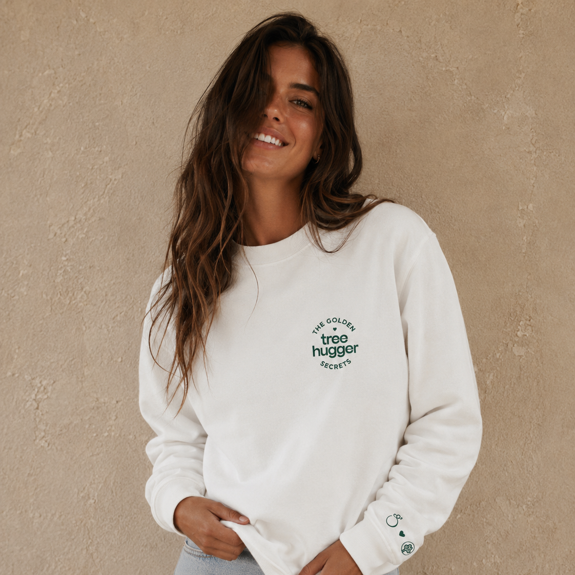 A woman with long brown hair, wearing THE GOLDEN SECRETS Tree Hugger Crewneck sustainable sweatshirt, smiles while standing in front of a beige textured wall.