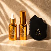 Three golden bottles from THE GOLDEN SECRETS Ultimate Glow Trio Kit and a black Gua Sha tool featuring gold logos are artfully arranged on a beige stone surface, bathed in soft sunlight and shadows to perfectly embody the radiant skin experience.
